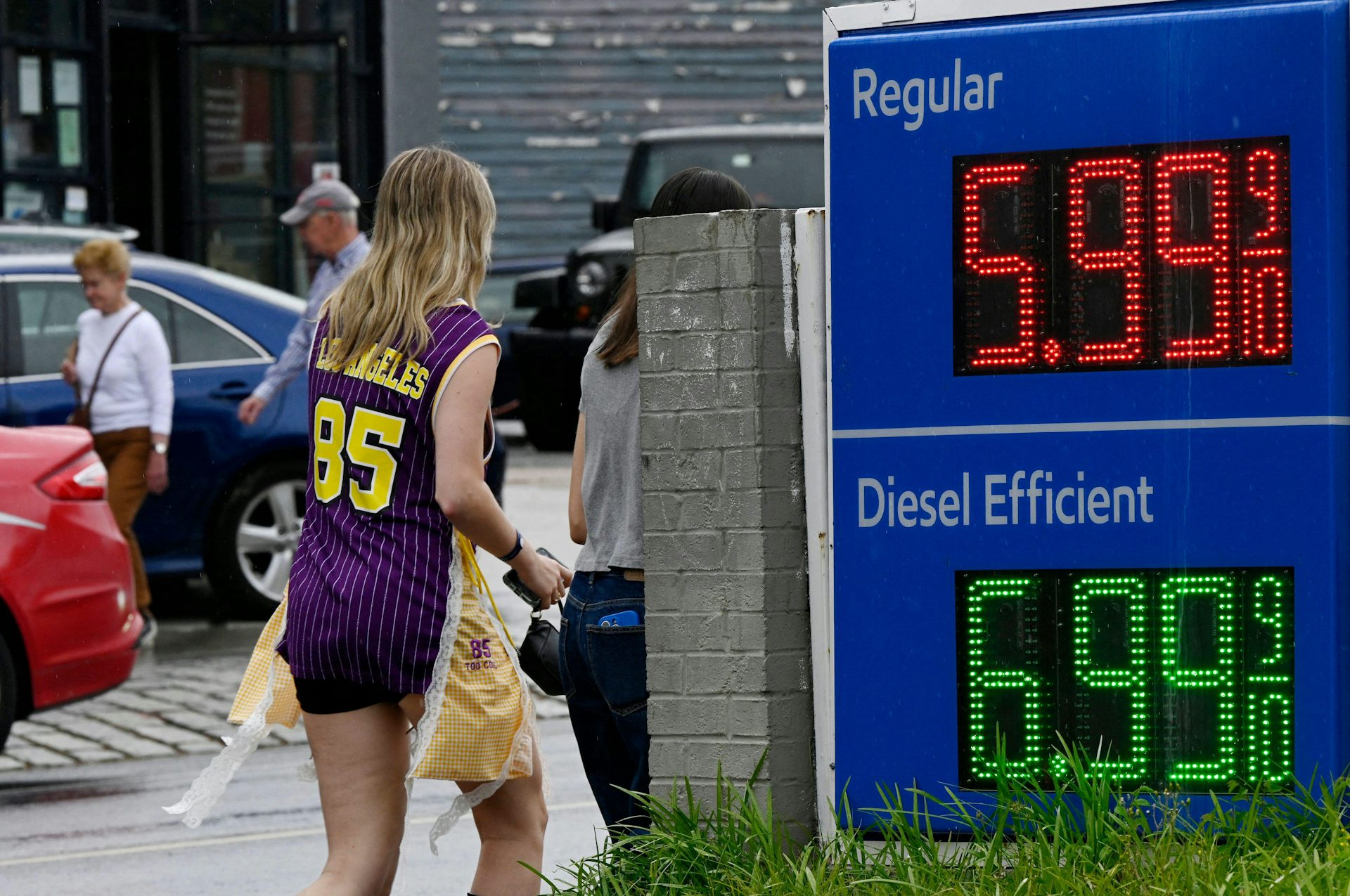 A woman in a football shirt walks away from a gasoline pump displaying high prices.