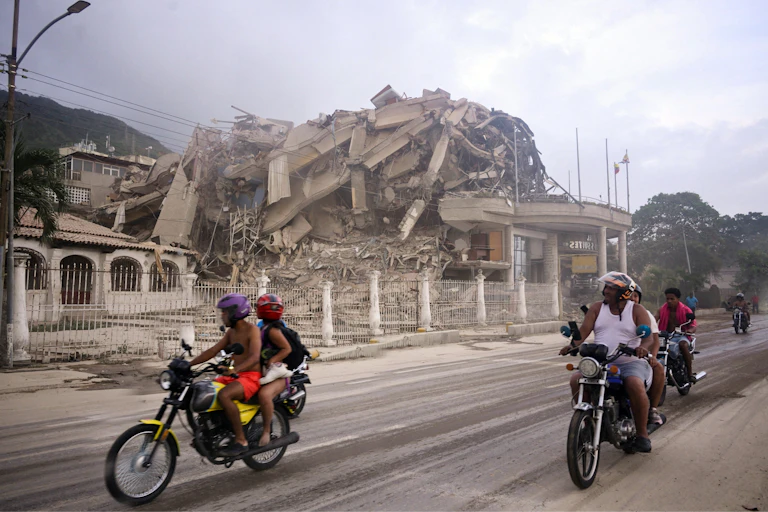 Varias motos circulan por una calzada con un edificio derruido al fondo