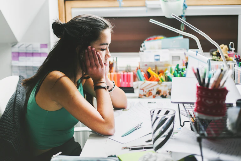 A teenager sits at a desk. Her head is in her hands.
