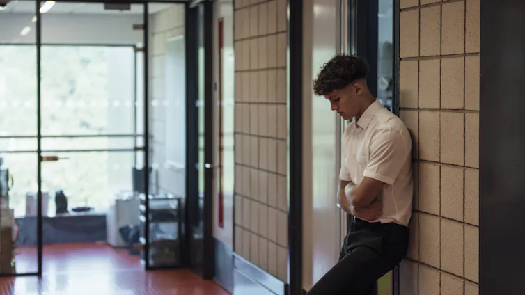 A side view of a teenage boy who is looking forlorn and sad after being sent out of the classroom. He has his arms folded and is looking down.