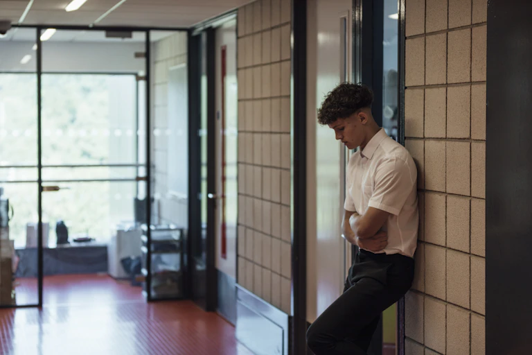 A side view of a teenage boy who is looking forlorn and sad after being sent out of the classroom. He has his arms folded and is looking down.