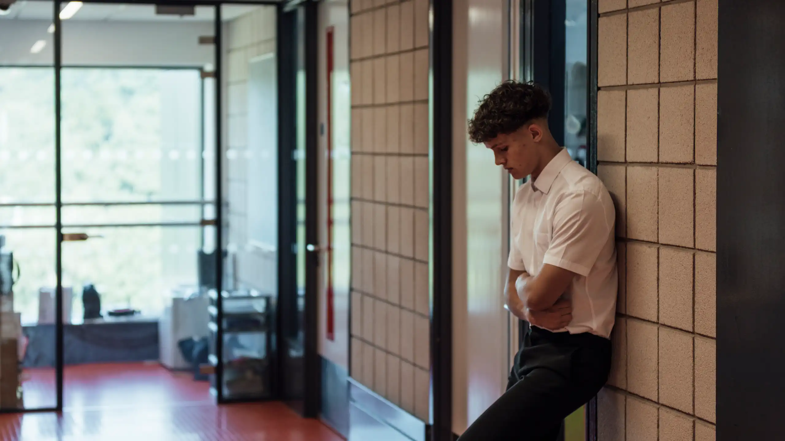 A side view of a teenage boy who is looking forlorn and sad after being sent out of the classroom. He has his arms folded and is looking down.