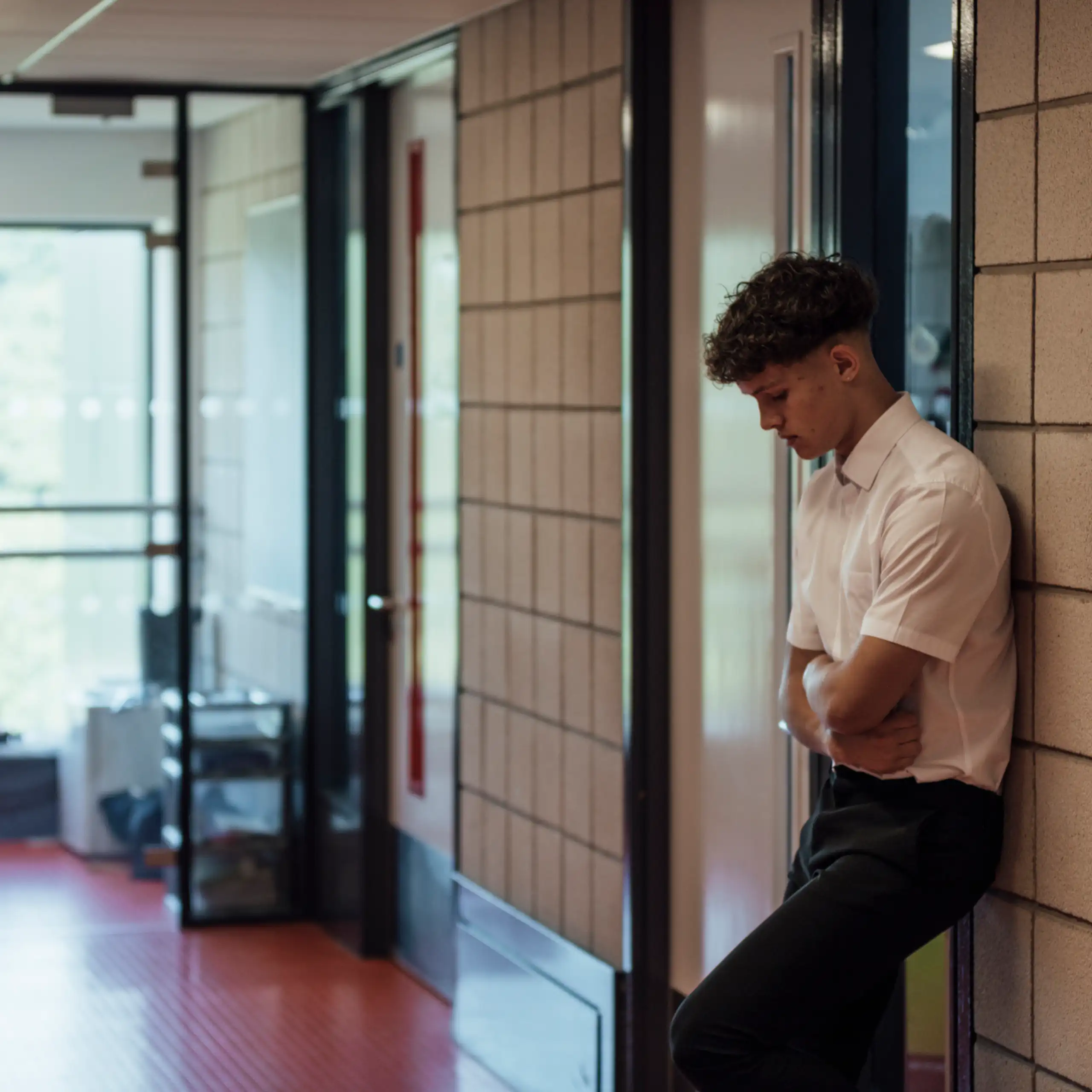 A side view of a teenage boy who is looking forlorn and sad after being sent out of the classroom. He has his arms folded and is looking down.