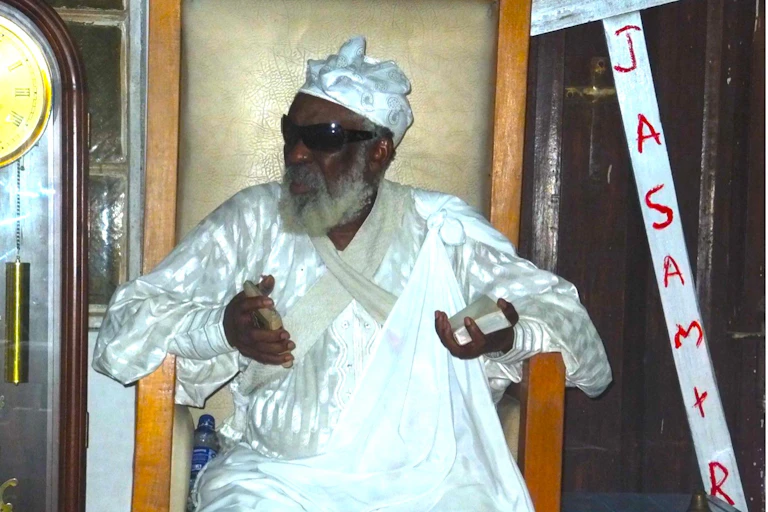 An African man with a grey beard wears a long white traditional garment and sunglasses as he sits in a chair holding a small book in either hand.