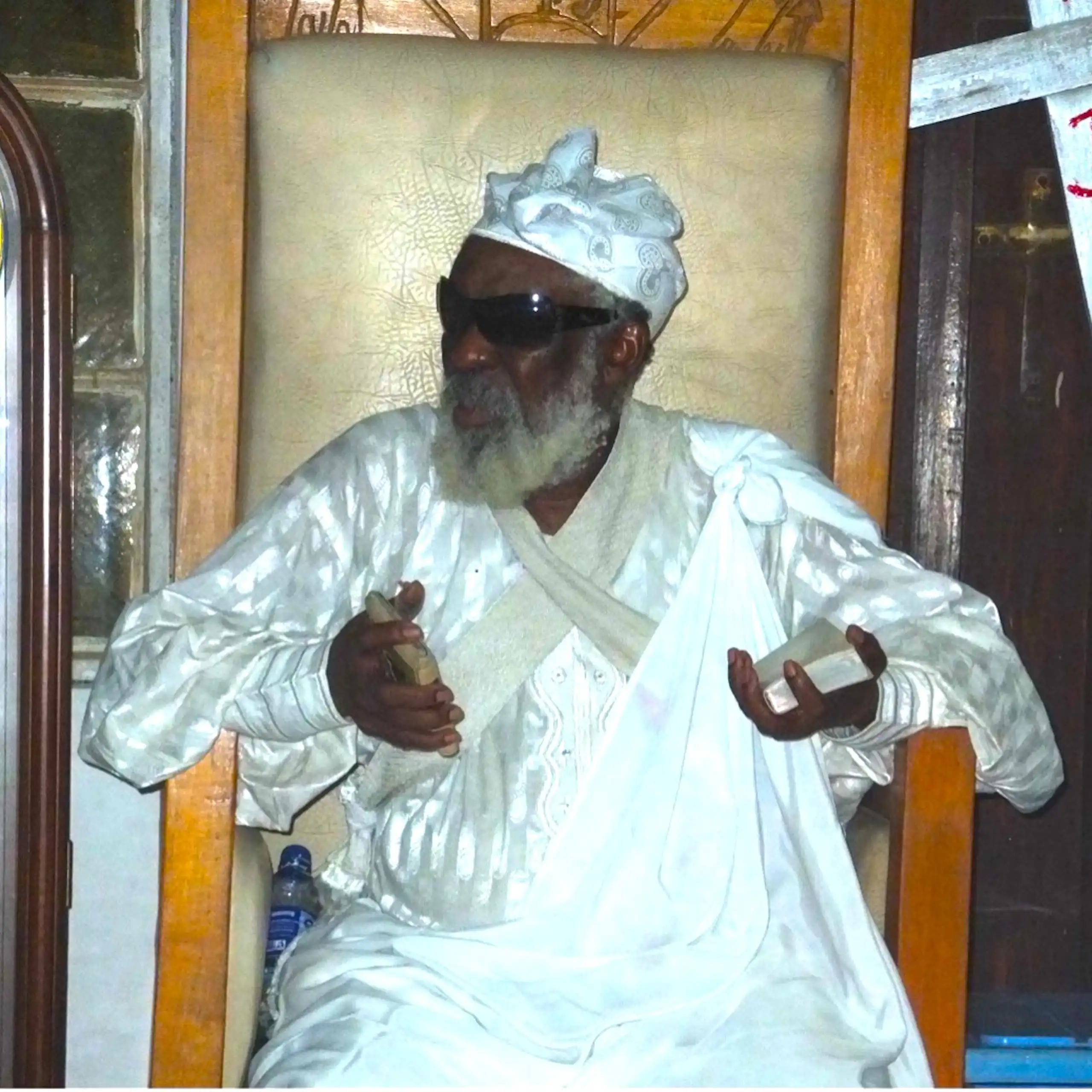 An African man with a grey beard wears a long white traditional garment and sunglasses as he sits in a chair holding a small book in either hand.