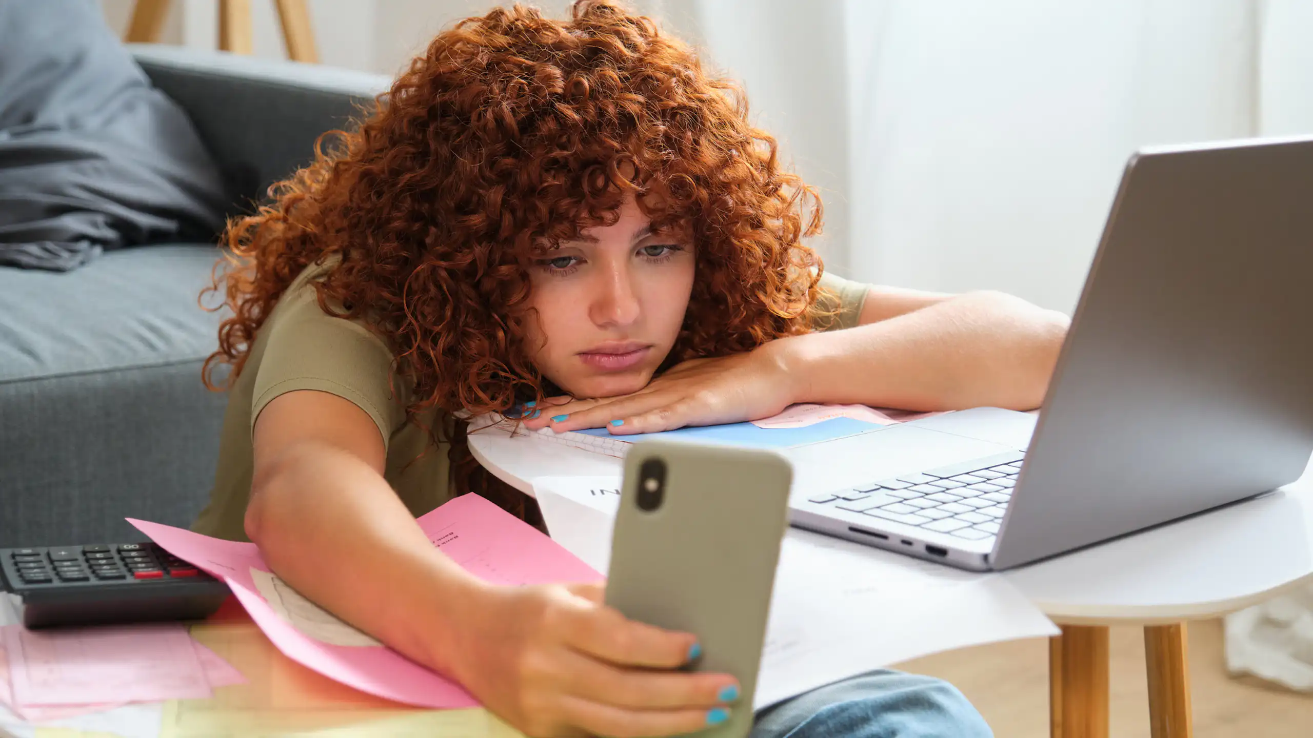 Une jeune femme regarde son téléphone, avachie devant son ordinateur portable, sur une table couverte de documents administratifs