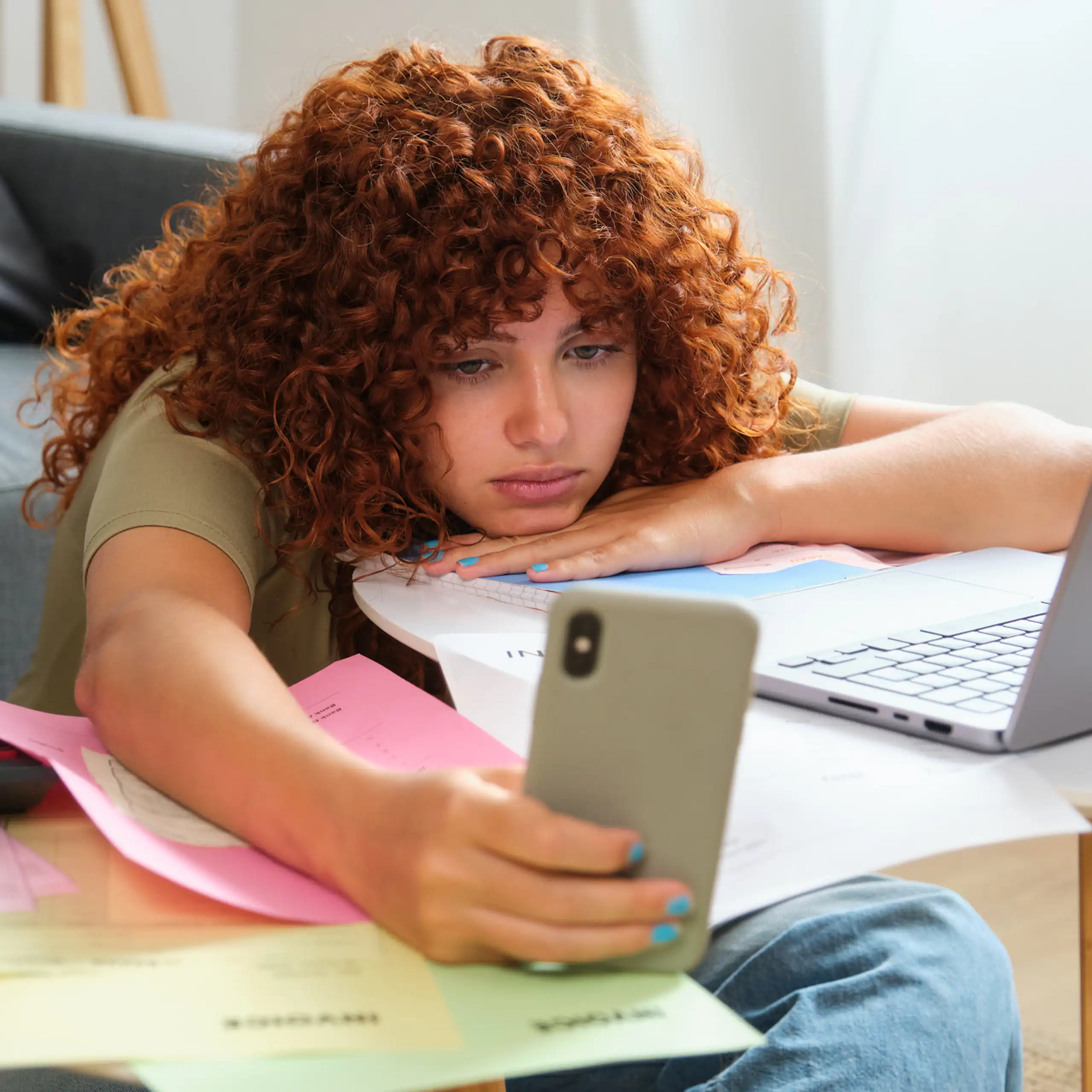 Une jeune femme regarde son téléphone, avachie devant son ordinateur portable, sur une table couverte de documents administratifs