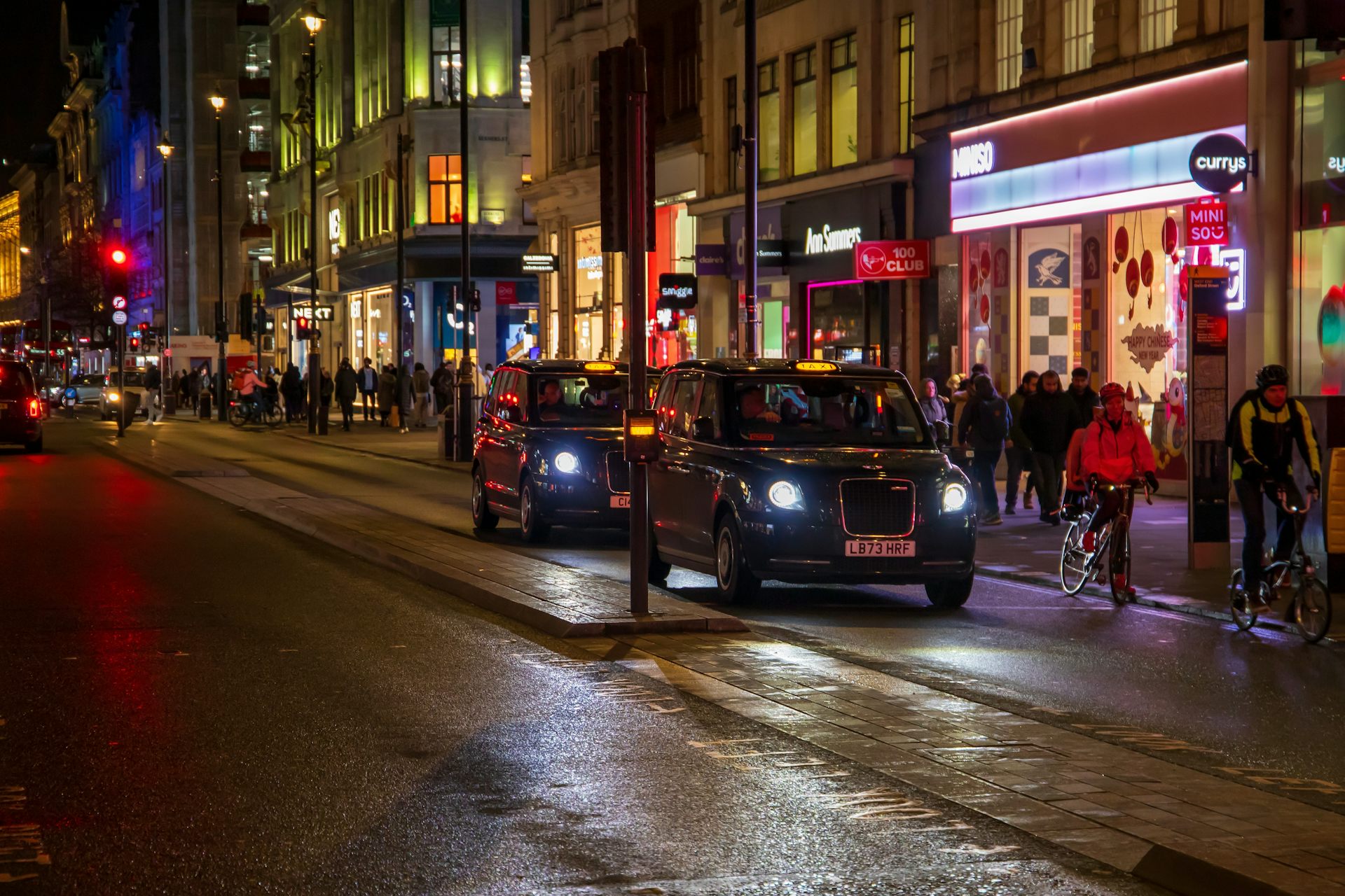 Cars driving on the street, people walking on the sidewalk with restaurants, retail stores and bars at night on the westside in London, England.