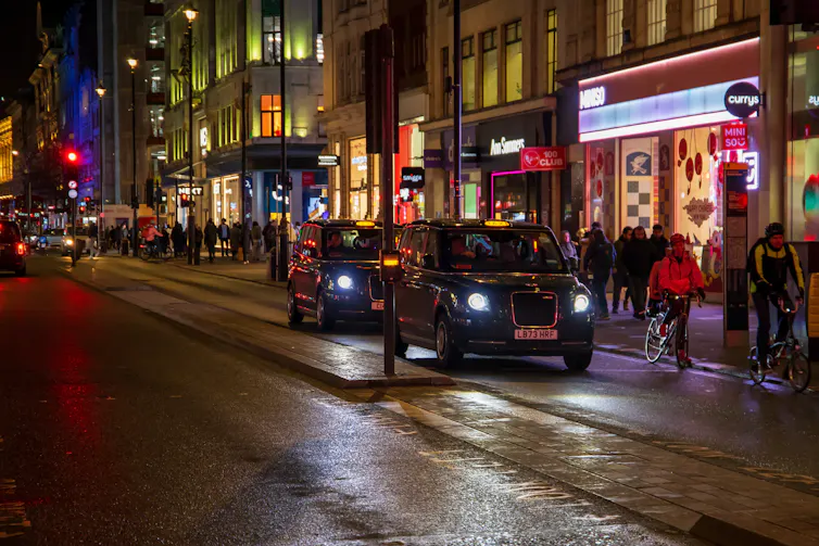 Cars driving on the street, people walking on the sidewalk with restaurants, retail stores and bars at night on the westside in London, England.
