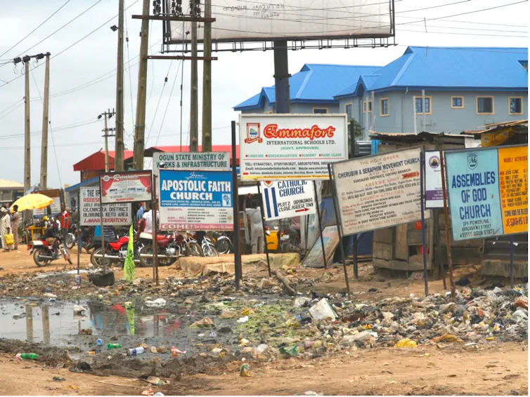 A dirt road with water pooled and lots of signboards alongside it, buildings in the distance.