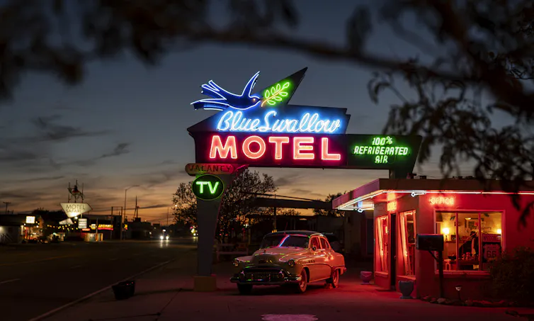Un motel de carretera al atardecer con un coche clásico aparcado en el aparcamiento y un gran letrero de neón azul y rosa que reza