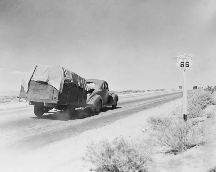 Fotografía en blanco y negro de un coche antiguo que arrastra un remolque con sus pertenencias por un tramo polvoriento de la carretera.
