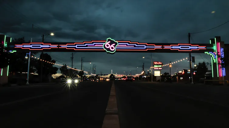 A road at dusk featuring neon signs, including a raised one spanning one side of the road to the other, featuring a neon version of a Route 66 highway marker.