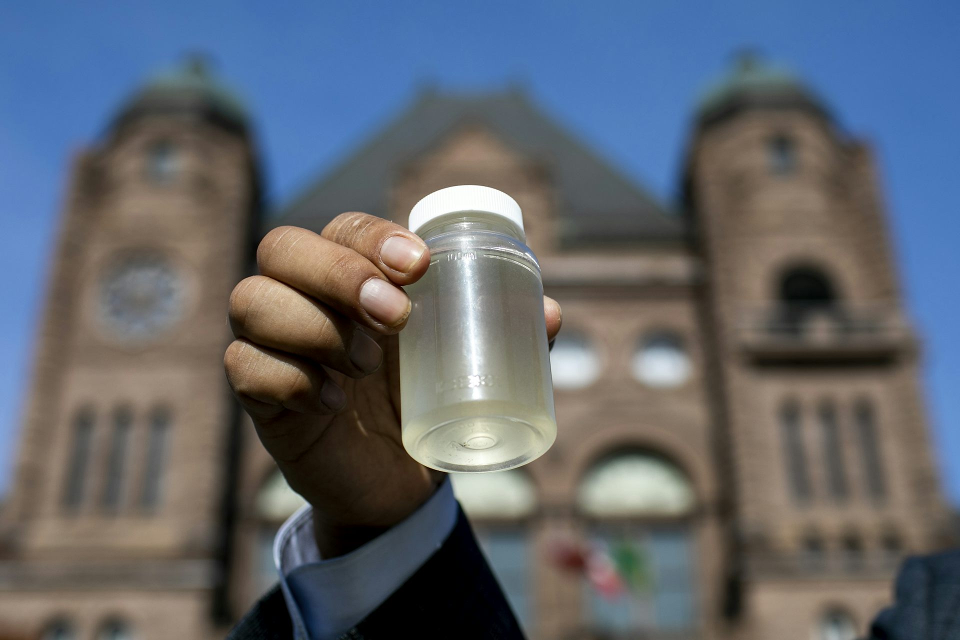 una mano sosteniendo una pequeña botella de agua contaminada frente a un gran edificio de piedra