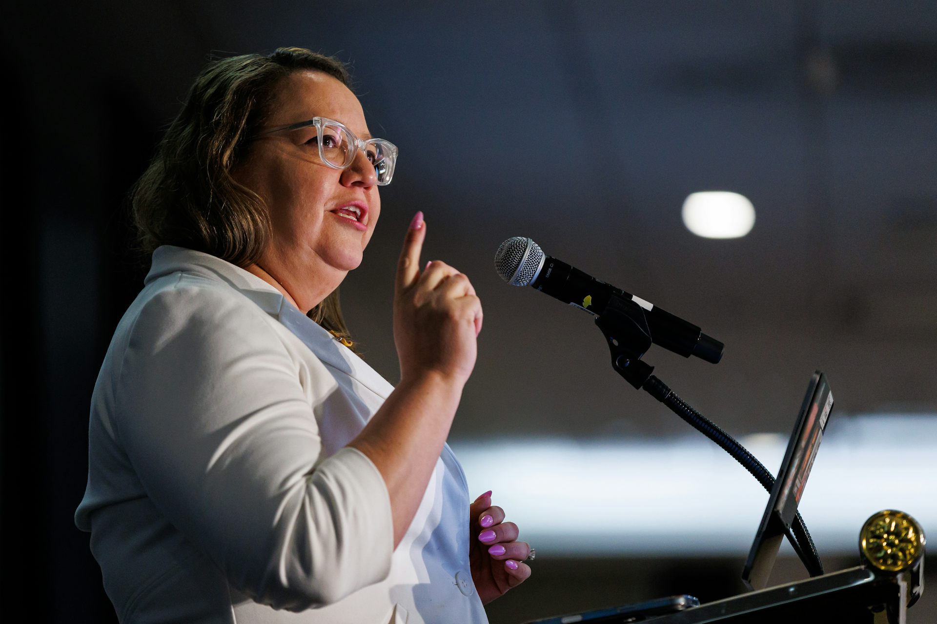 A woman wearing glasses and a white shirt gestures while speaking into a microphone