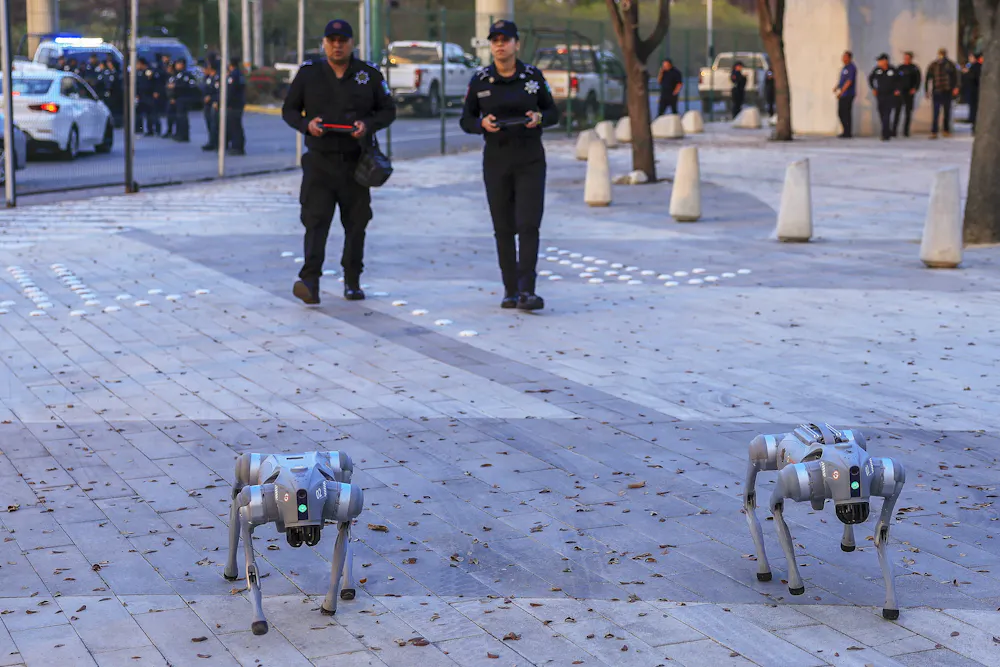 Image 1: two people wearing uniforms walk behind two robotic dogs