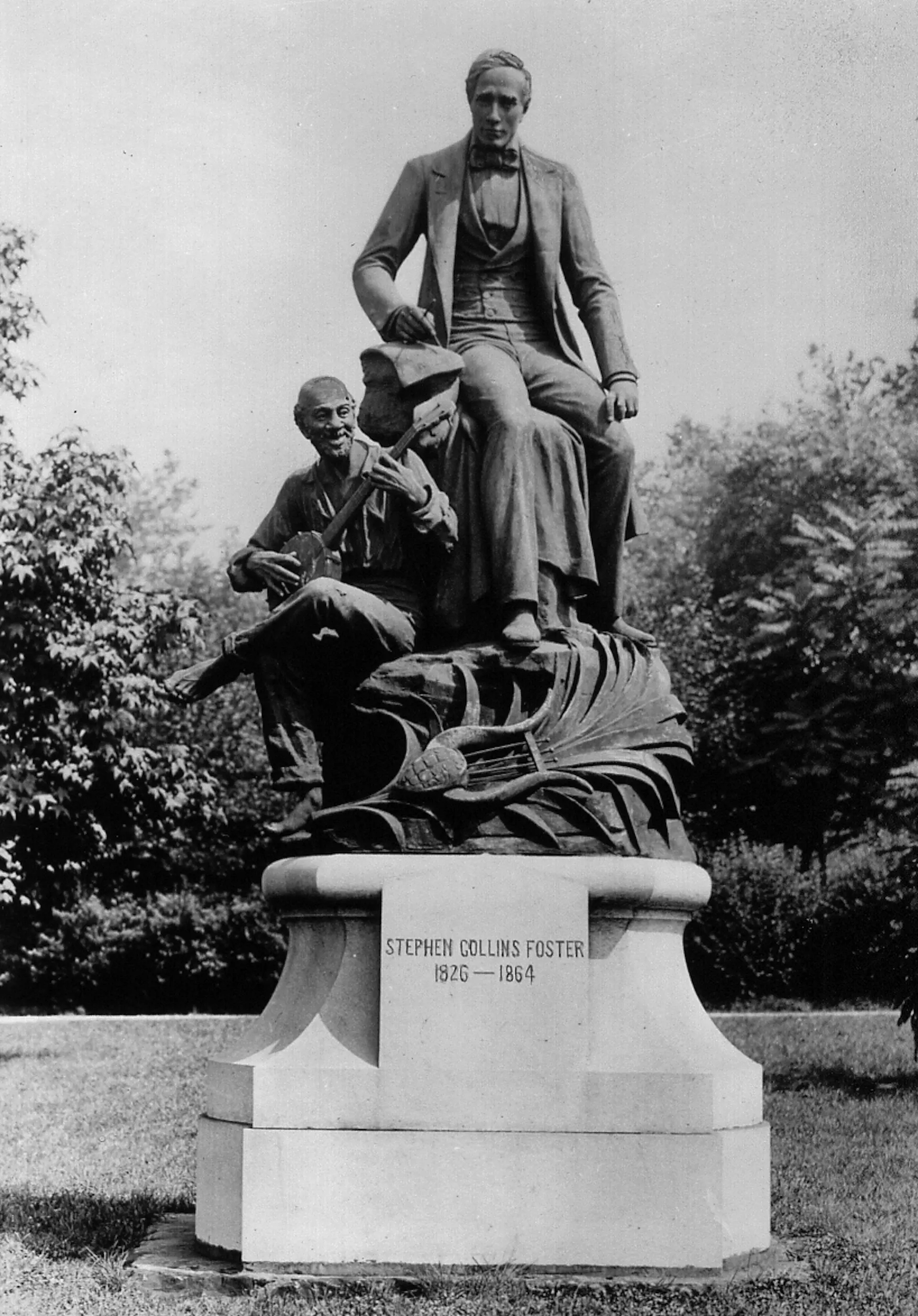 Black and white photo of a statue depicting a white man wearing a suit while a Black man sits by his sides with his legs crossed, playing a banjo.