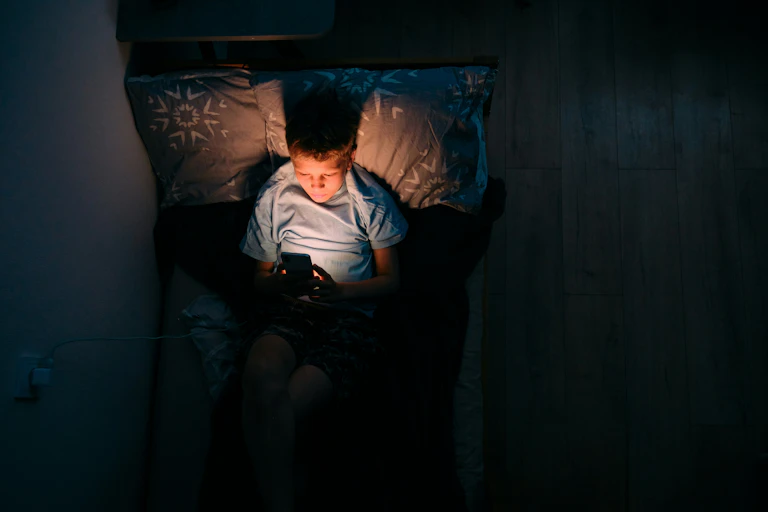 A boy laying on his bed and looking at his smart phone in a dark room.