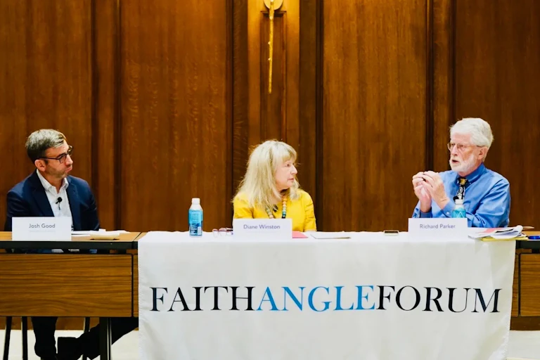 Two men and a woman (in the center) are at the front of a room behind a brown table. A banner fronting the table reads Faith Angle Forum.