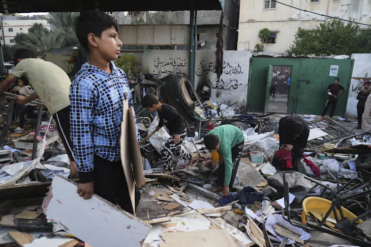 A group of boys stand amid the rubble of a destroyed building, picking up pieces