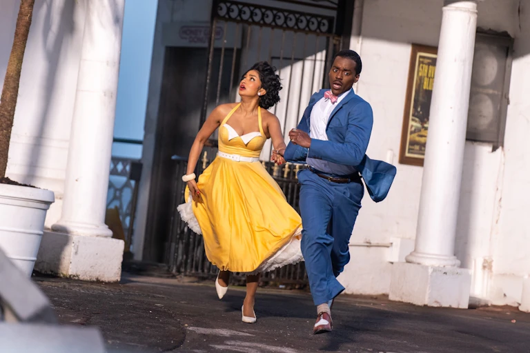 Production image: The Doctor and his assistant Belinda Chandra run away from a building.