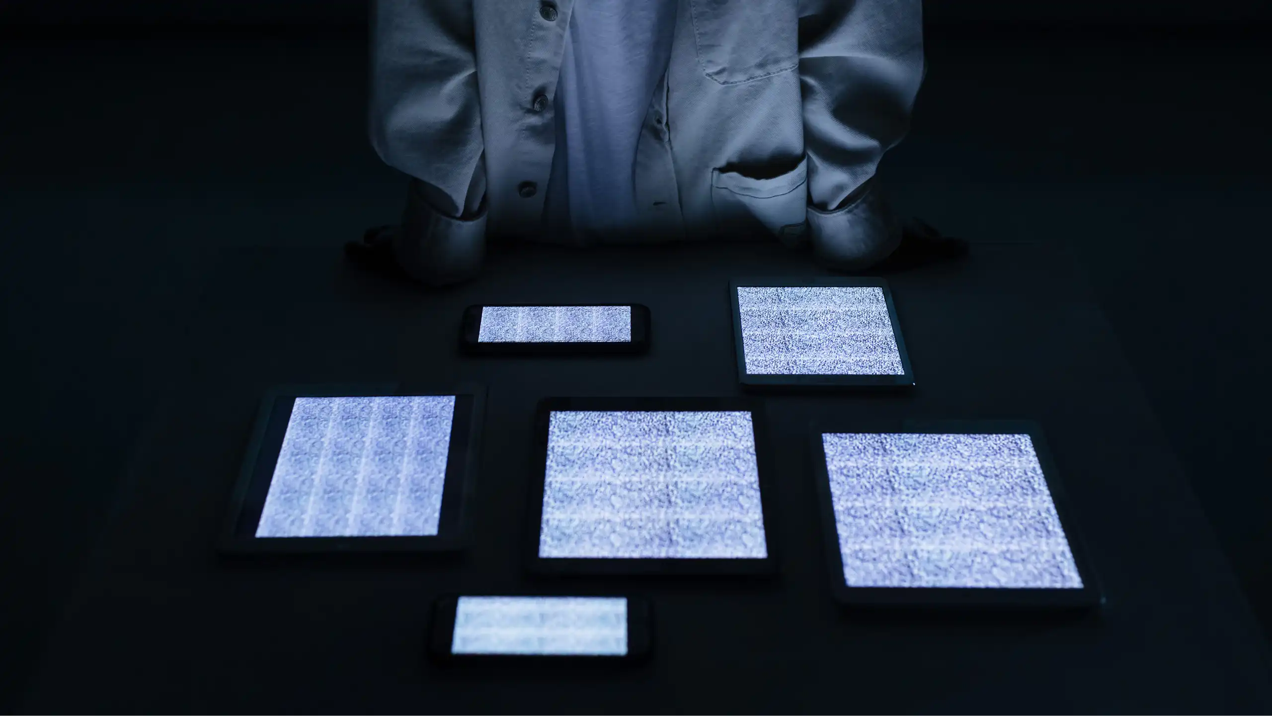A man in a white top stands behind a table full of screens with static on them