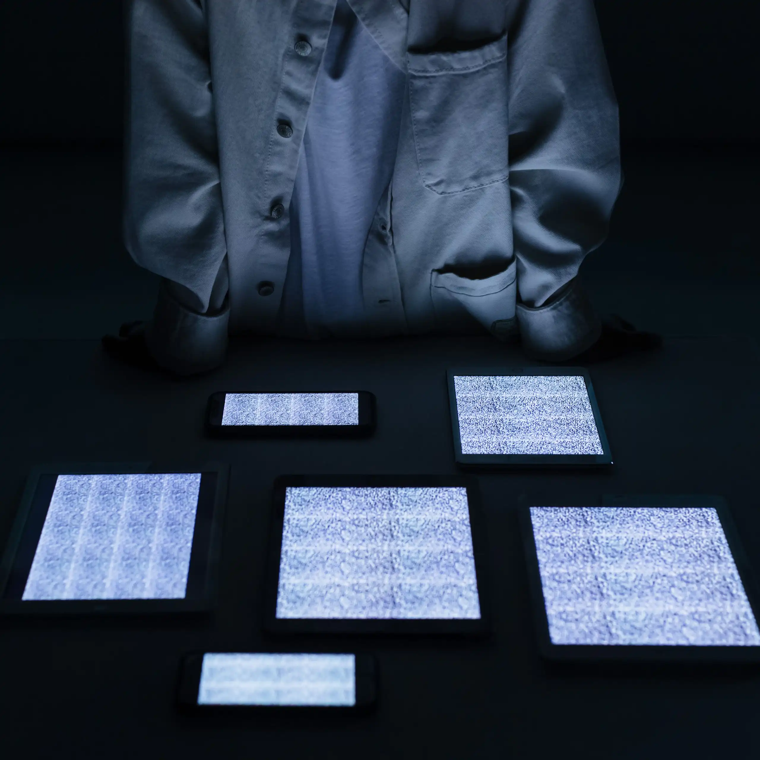 A man in a white top stands behind a table full of screens with static on them