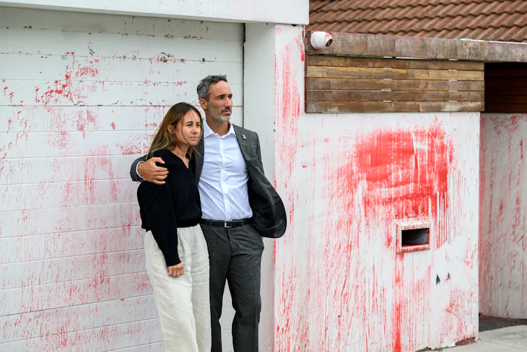 A man and a woman stand outside a white house graffitied with red spray paint