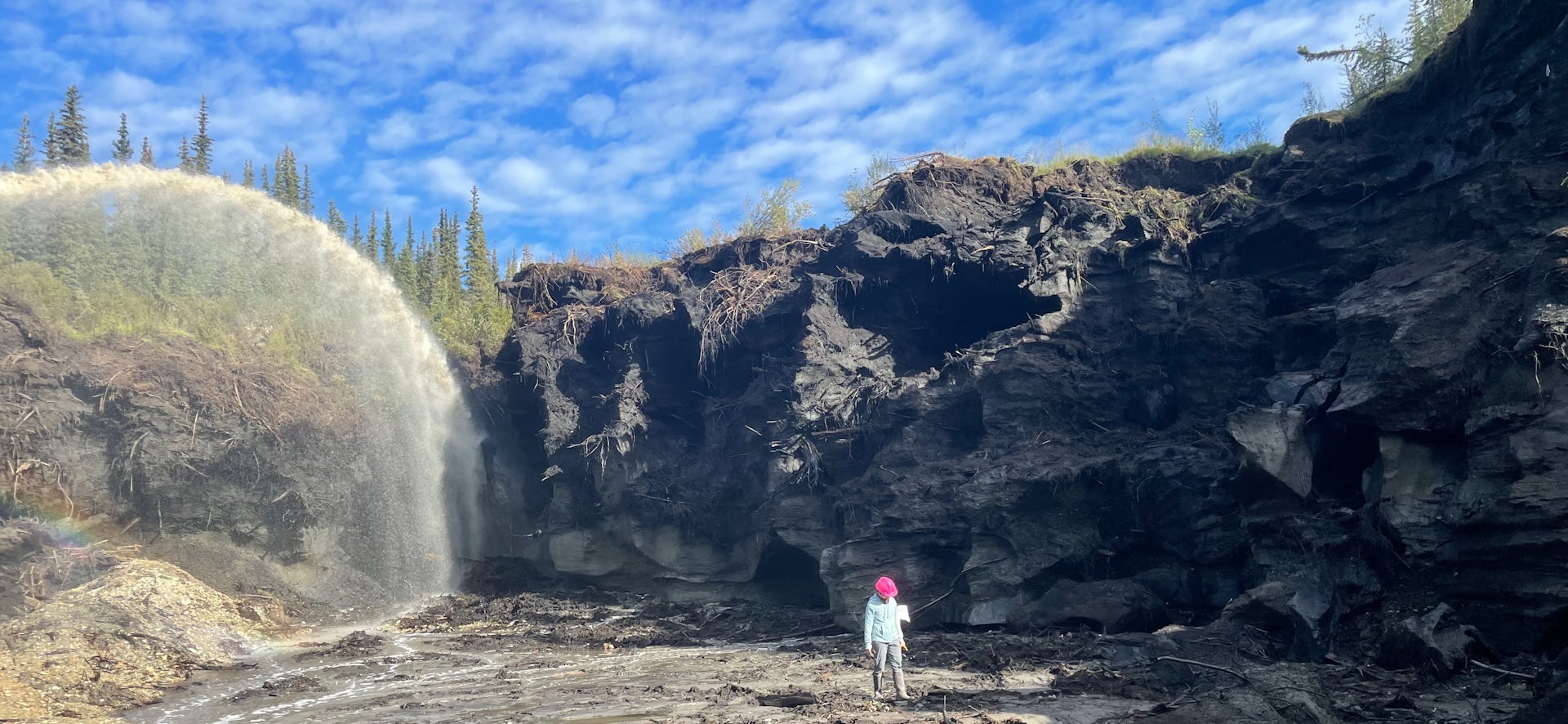 Un hombre con sombrero rojo camina frente a un acantilado y una gran corriente de agua, con un cielo azul detrás