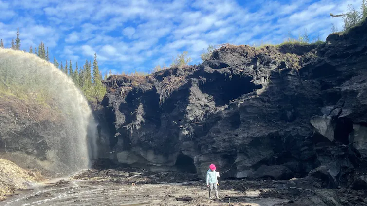 La memoria profunda de la Tierra se está derritiendo con el permafrost ártico, degradando el registro de nuestro mundo antiguo 2 Un hombre con sombrero rojo camina frente a un acantilado y una gran corriente de agua, con un cielo azul detrás