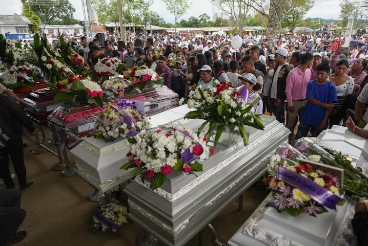 A crowd of people around coffins covered wtih flowers.