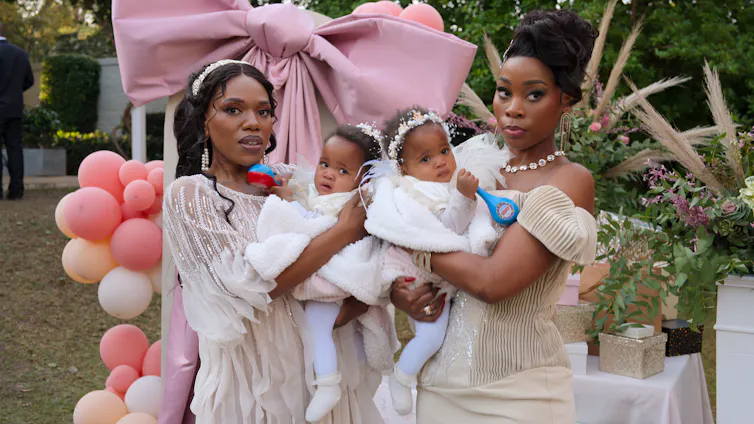 Two smartly dressed African women at a garden party with balloons, each holding a baby.