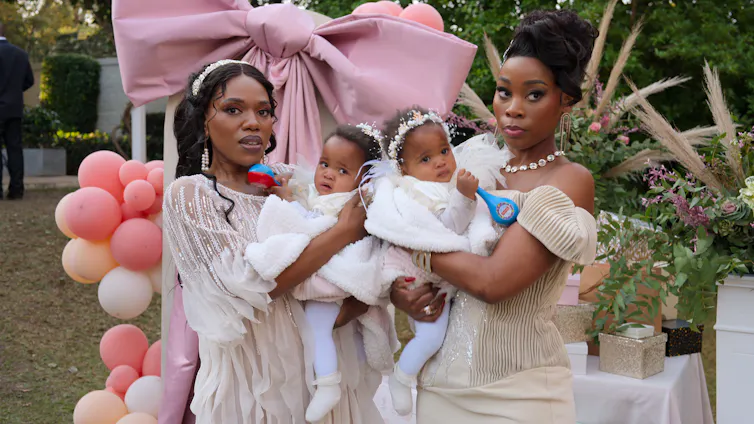 Two smartly dressed African women at a garden party with balloons, each holding a baby.