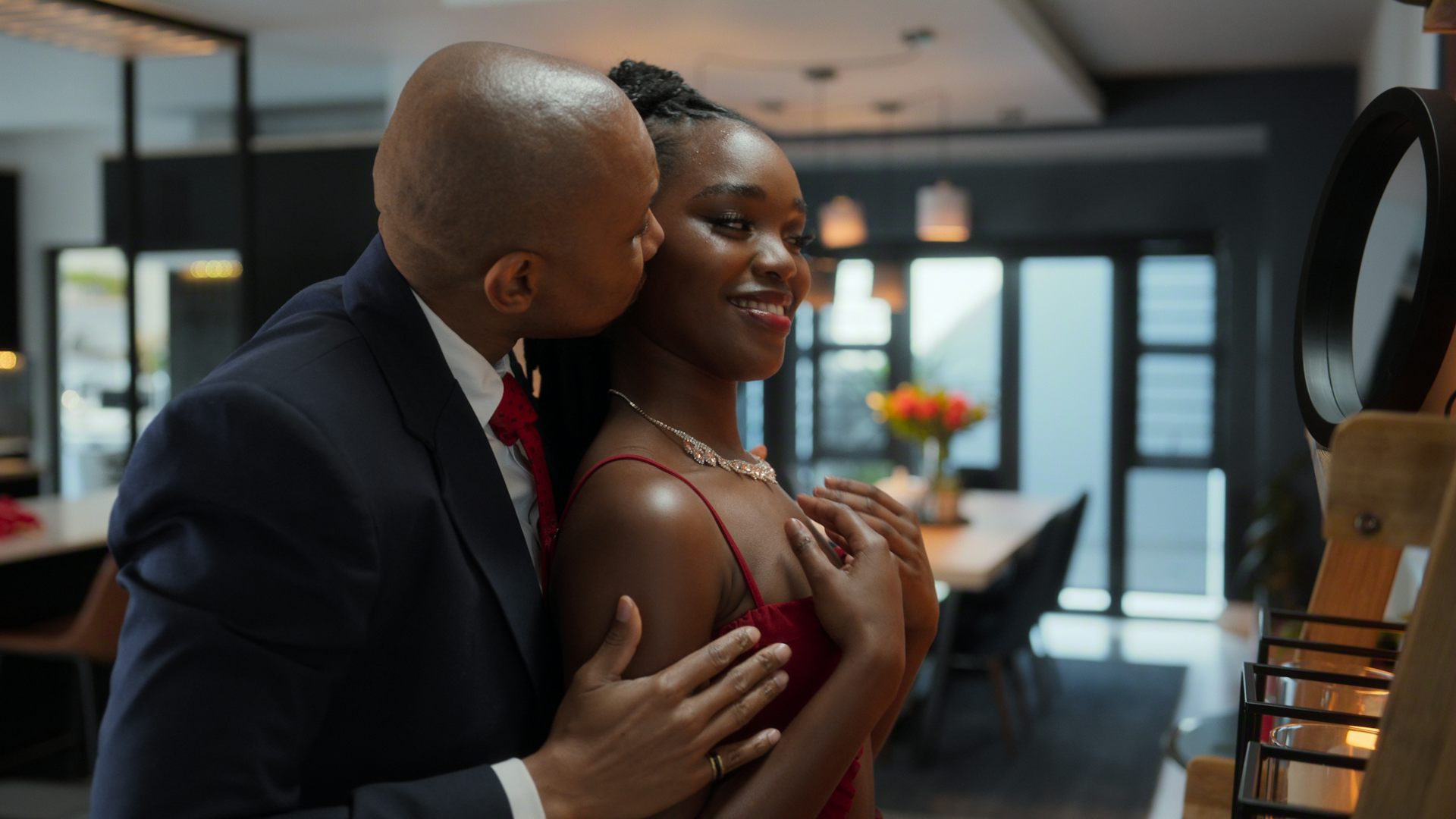 An African man in a suit kisses a young African woman from behind as she smiles into a mirror.