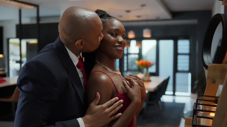An African man in a suit kisses a young African woman from behind as she smiles into a mirror.