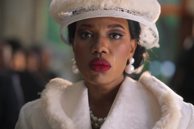 An African woman smartly dressed stares straight ahead wearing bright red lipstick and a white hat, tears stains down her face.