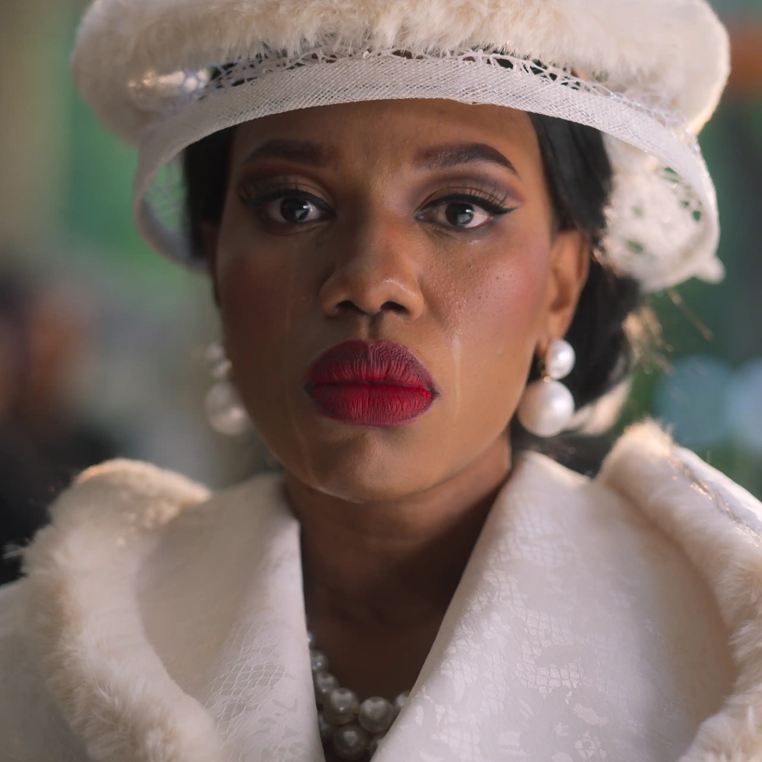 An African woman smartly dressed stares straight ahead wearing bright red lipstick and a white hat, tears stains down her face.