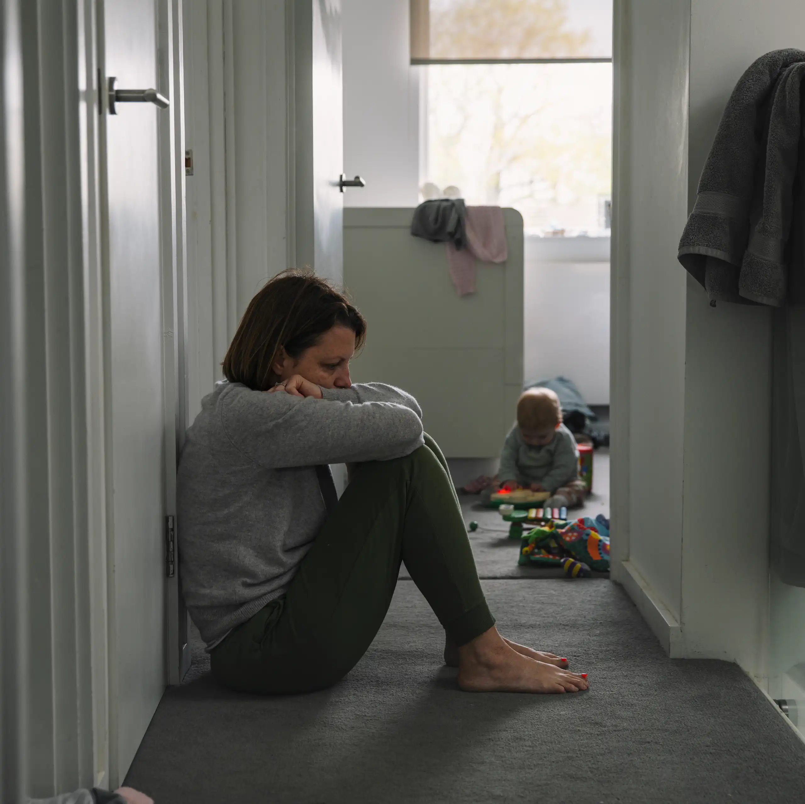 A woman sits in a hallway while an infant plays in the room beyond her.