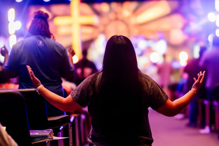 A long-haired person from behind, hands raised, bright light in front of them.