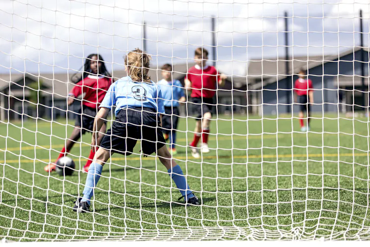 View behind a soccer goal, three children in red uniform approaching a child in a blue uniform as their teammate is also approaching