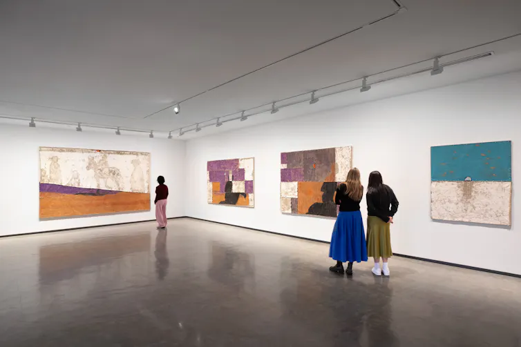 A large white gallery. Three people look at pastel paintings.