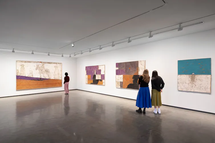 A large white gallery. Three people look at pastel paintings.