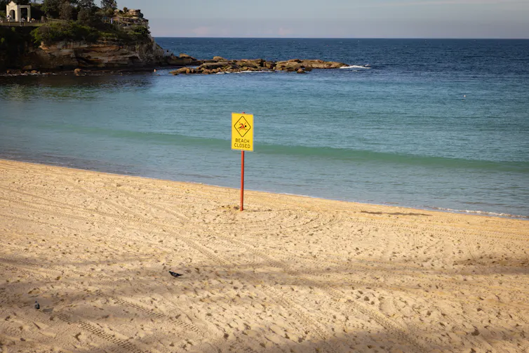 sign saying beach closed, empty beach and calm sea behind.