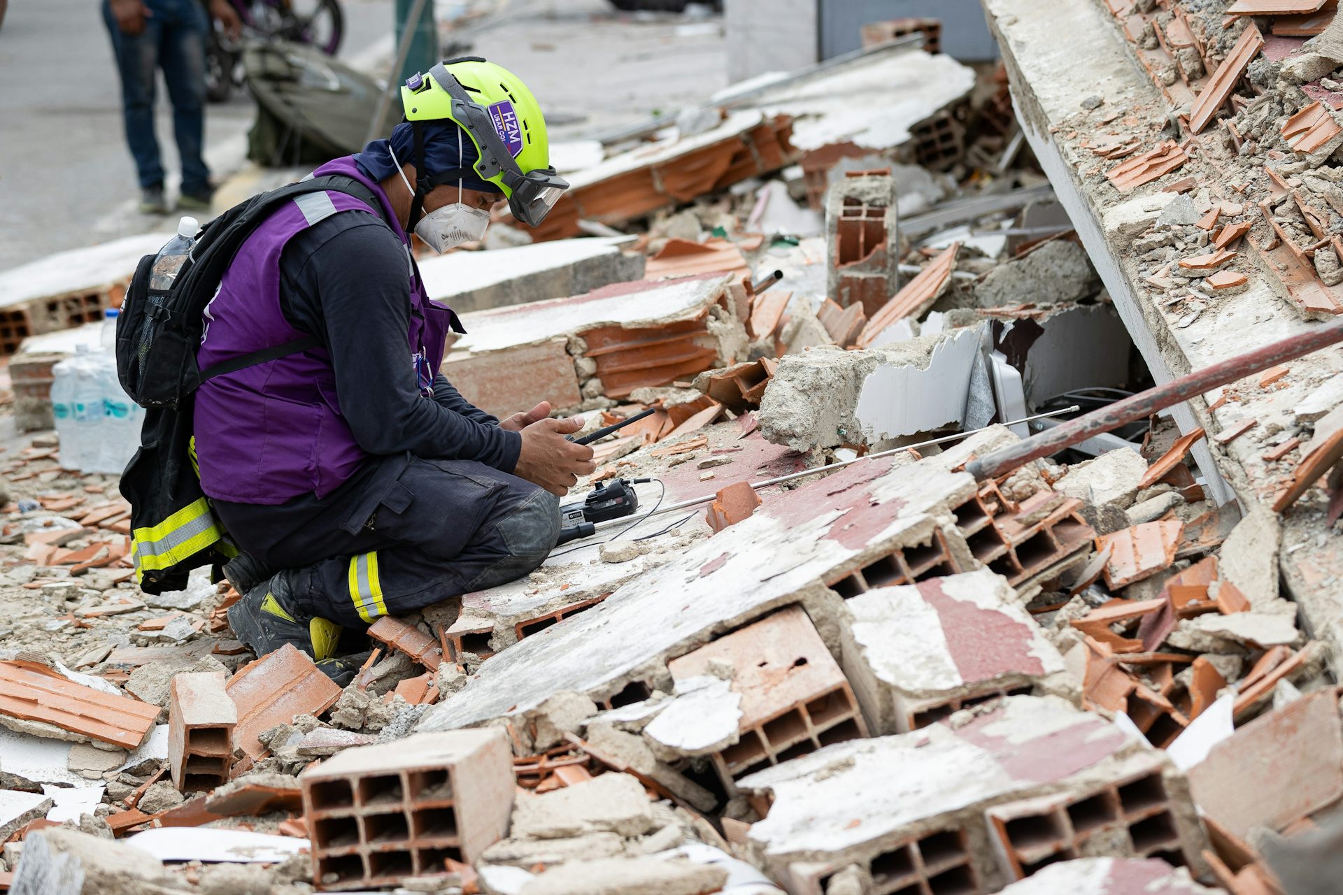 Rescue worker in purple tabard and hi-vis helmet crouches amid rubble