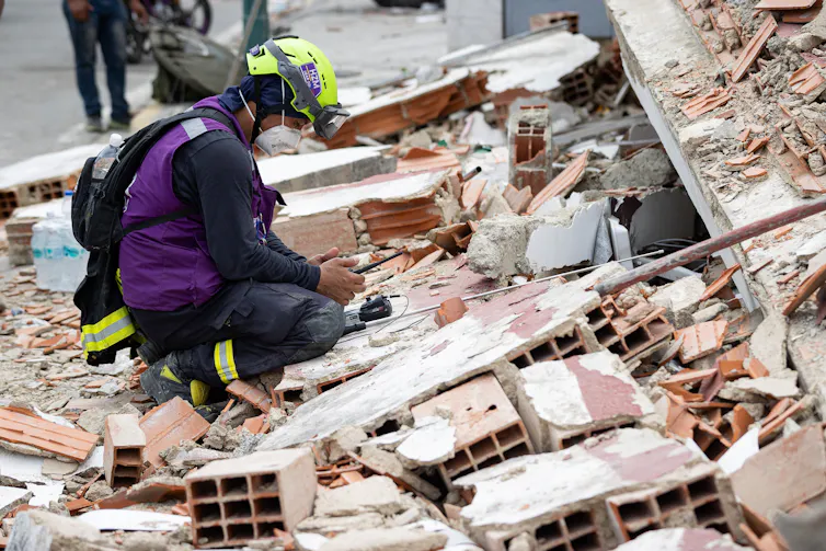 Rescue worker in purple tabard and hi-vis helmet crouches amid rubble