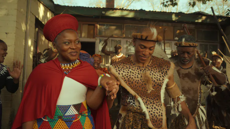 A customary African wedding in traditional attire, a smiling bride and groom walk holding hands.