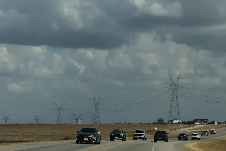 A line of tall metal towers holding wires stretches into the distance over a highway.