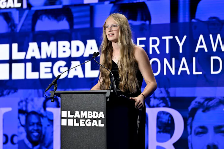 A young woman with long light brown hair stands at a podium that says 'Lamba Legal' with a blue backdrop behind her that has the same words.