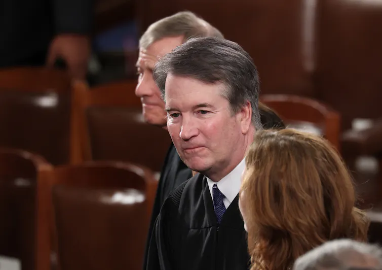 The face of a man with dark brown hair, who is wearing a black robe, is seen between two other people.