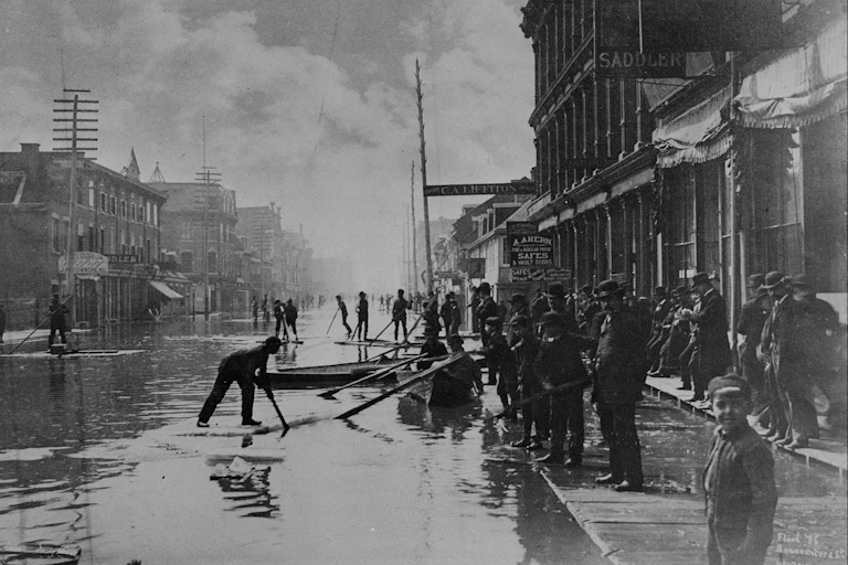 a black and white photo of people standing on the side of a flooded road lined with brick buildings.